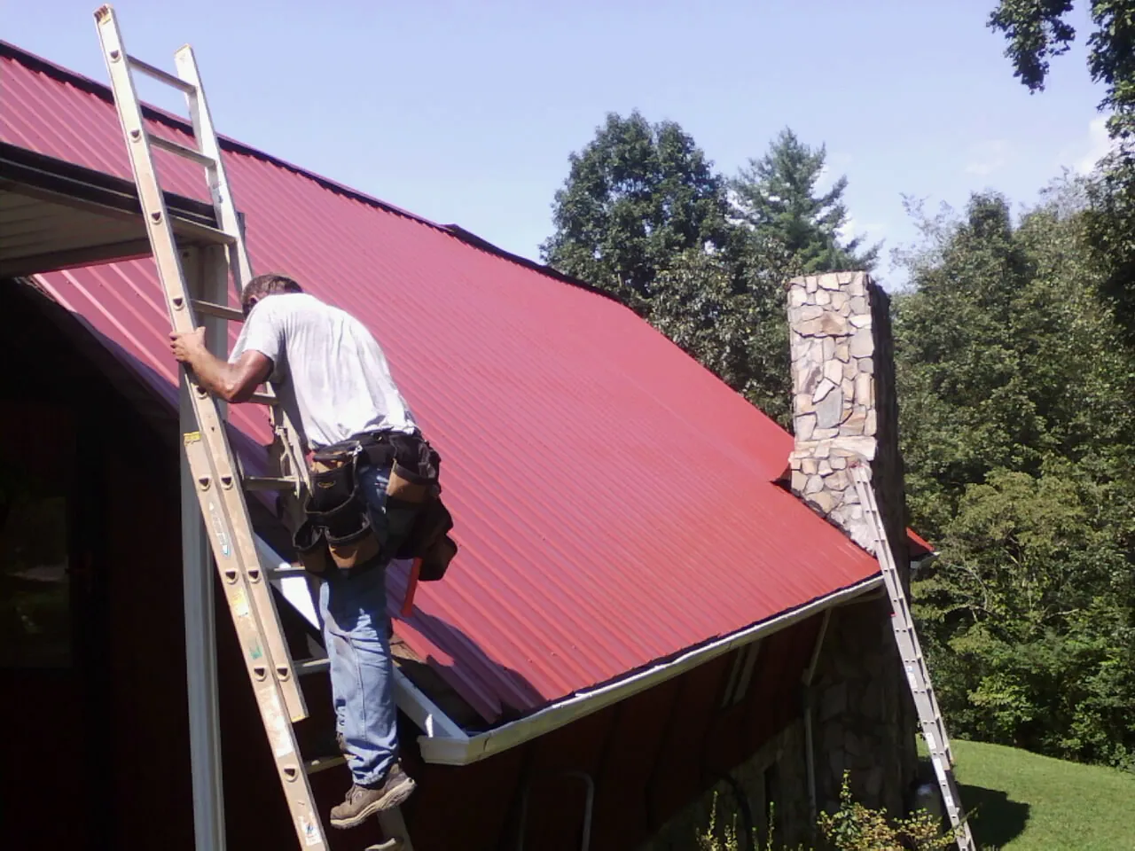 Roofer on a ladder beside a red metal roof and stone chimney — Soffit & Fascia Repair by Roofing Brothers in Loon Lake